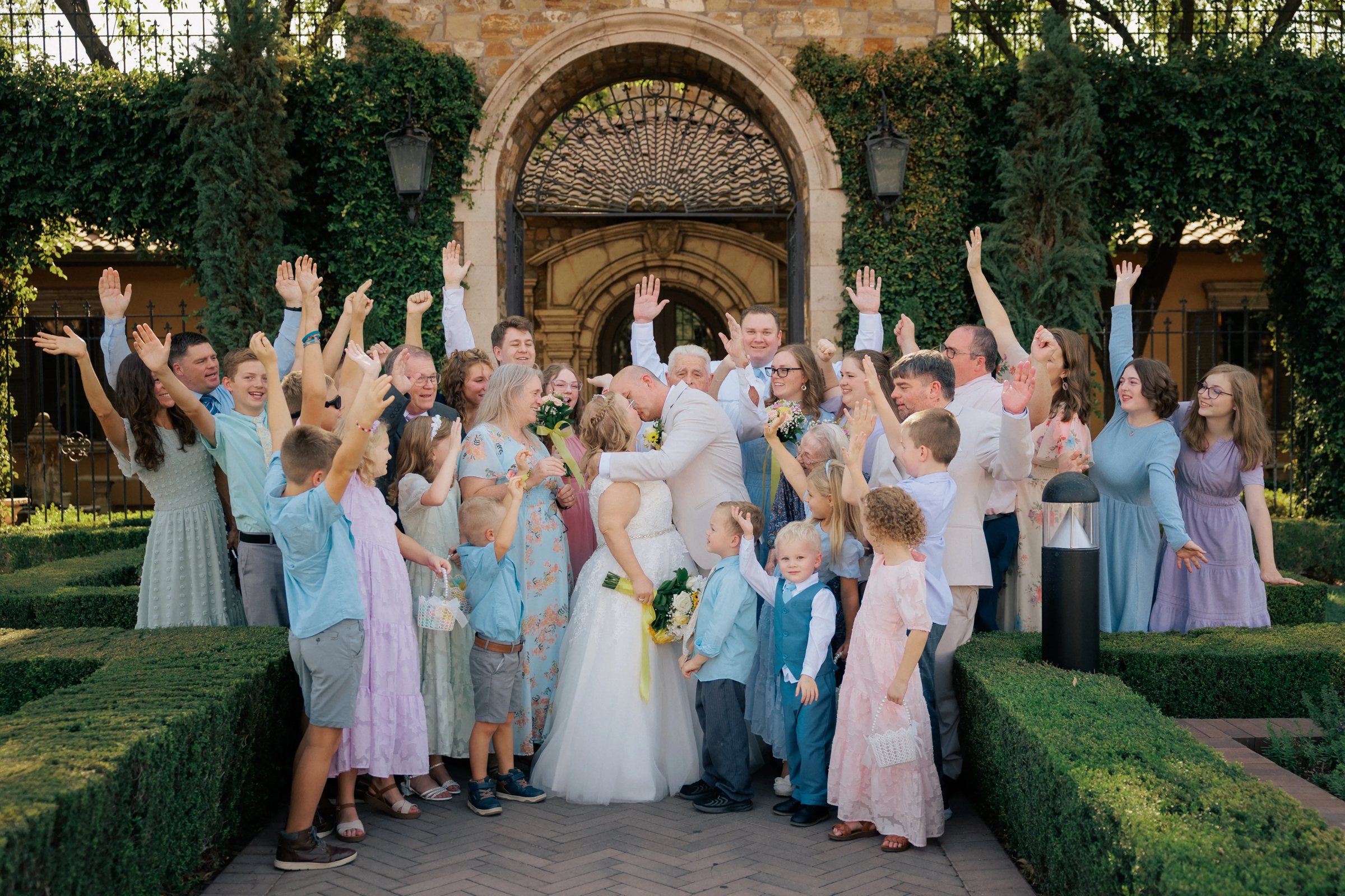 Bride and groom kiss in front of the arched iron gates at Villa Siena in Gilbert, Arizona, surrounded by cheering family