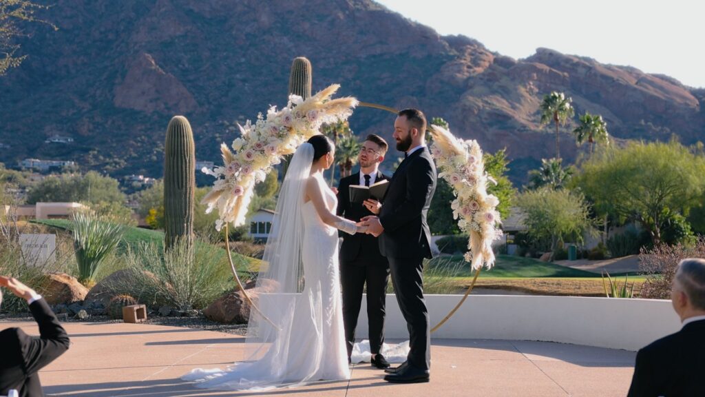 Couple exchanging vows under a floral hoop on the Mountain Shadows Lawn, with Camelback Mountain behind